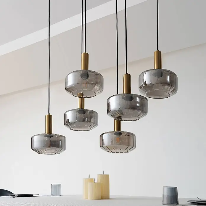 Lindby Pendelleuchte Hildur - alu / grau / zink, rauchgrau, messing, E27 - Sechs Pendelleuchten mit Rauchglas-Schirmen und Messingakzenten über einem Esstisch mit Kerzen.