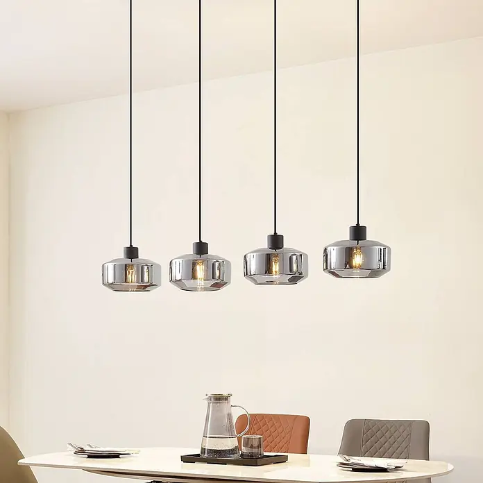 Lindby Pendelleuchte in rauchgrau transparent, schwarz matt, Hängelampe -  - Vier moderne Pendelleuchten mit rauchgrauen Glasschirmen und schwarzen Fassungen hängen beleuchtet über einem Esstisch.