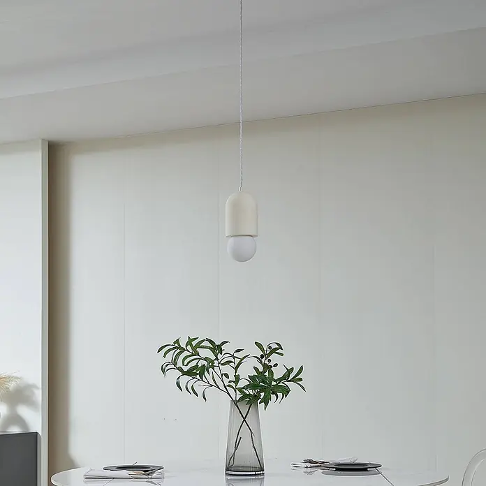 Lindby Pendelleuchte Nielson - creme / amber, beige, weiß, E27 - Minimalistische weiße Pendelleuchte mit pillenförmigem Gehäuse und runder Glühbirne über einem Esstisch mit Pflanze.