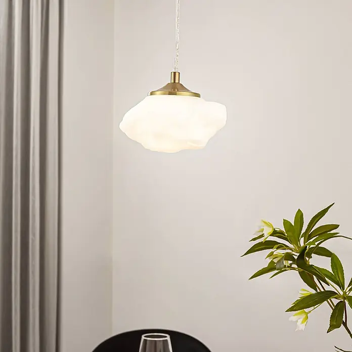 Lindby Pendelleuchte Piatra - L x B x H: 15,5 x 15,5 x 8,5 cm, weiß / opal, weiß, messing, G9 - Eine Pendelleuchte mit weißem, wolkenförmigem Glasschirm und Messingfassung, hängt in einem Raum über einem Tisch.