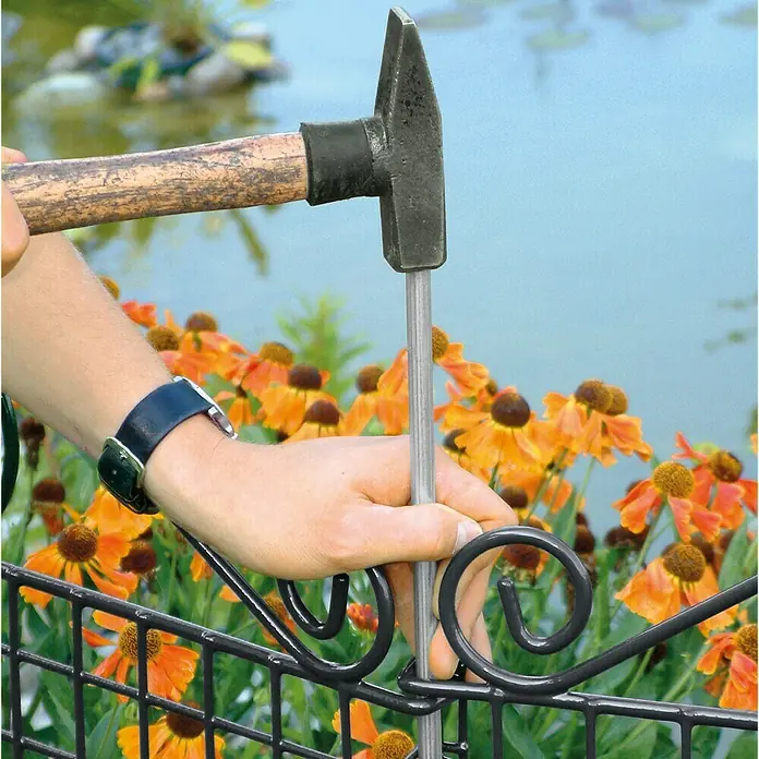 Bellissa Zusatzbodenanker - Länge: 65 cm, Feuerverzinkt - Ein Mann befestigt mit einem Hammer einen Zaunpfahl an einem dekorativen Gartenzaun, umgeben von bunten Blumen.