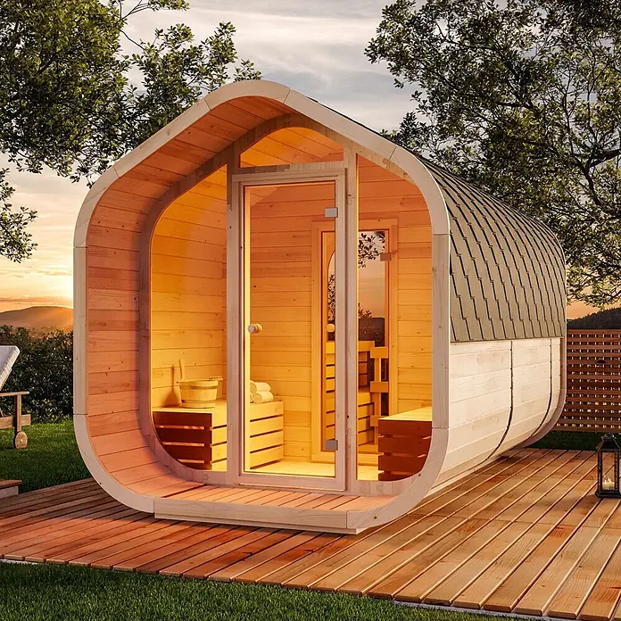 FinnTherm Fasssauna ScandiCube Wave Deluxe - Holz, L x B x H: 406 x 237 x 249 cm - Eine moderne Fasssauna aus hellem Holz mit dunklem Dach und Glastür, auf einer Holzterrasse bei Sonnenuntergang.
