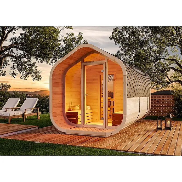 FinnTherm Fasssauna ScandiCube Wave Deluxe - Holz, L x B x H: 406 x 237 x 249 cm - Eine moderne, helle Fasssauna aus Holz mit Schindeldach, die bei Sonnenuntergang in einem Garten einladend leuchtet.