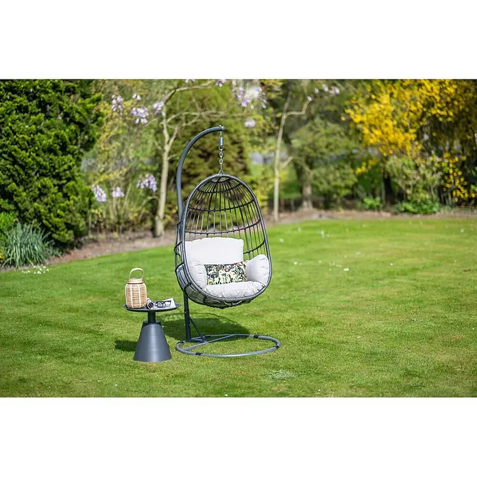 Garden Impressions Hängesessel Fargo - L x B x H: 92 x 79 x 198 cm, Metall, Schwarz/Grau - Dunkelgrauer geflochtener Hängekorbsessel mit hellen Kissen und Ständer. Ein Beistelltisch mit Laterne steht daneben.
