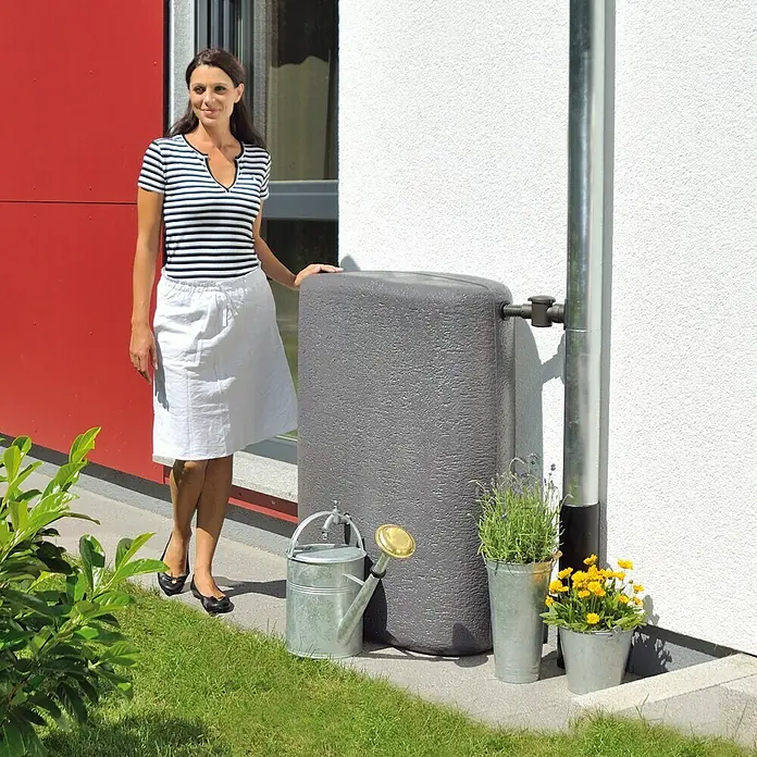 Garantia Classic Line Regenspeicher Terranova - 275 l, Graphit - Frau neben einer grauen Regentonnen mit Gießkanne und Blumen. Eine effiziente und nachhaltige Lösung zur Wasserspeicherung für den Garten.