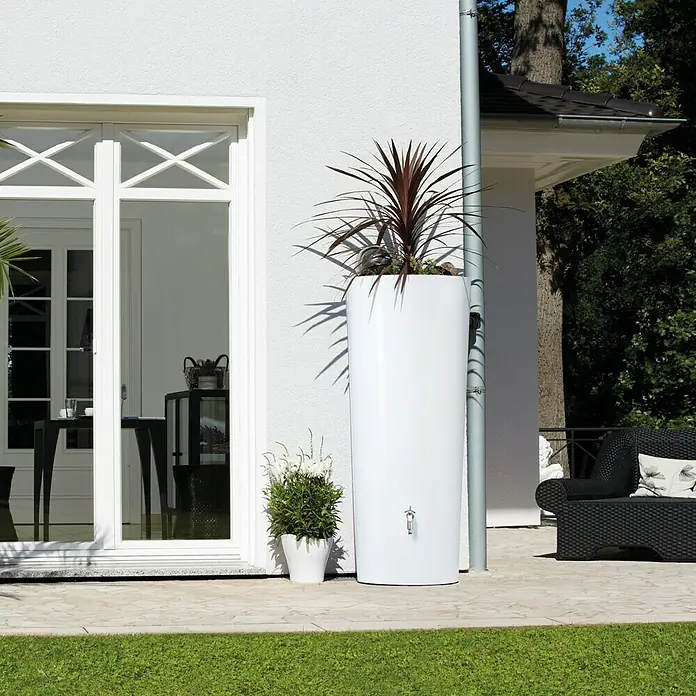 Garantia Modern Line Regenspeicher Color 2 in 1 - 350 l, Coco - Eine weiße Regentonnen-Pflanzgefäß-Kombination steht vor einem Haus, bepflanzt mit einer roten Pflanze. Eine kleine Pflanze steht daneben.
