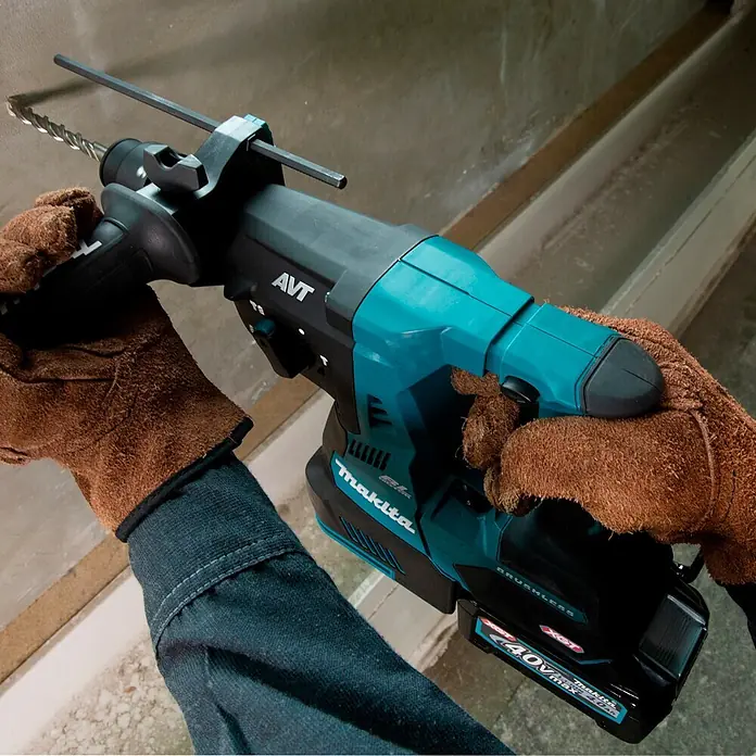 Makita XGT 40V max. Martillo combinado de batería HR004GZ - 40 V, Sin batería, 2,9 J - Manos enguantadas operan un taladro percutor Makita turquesa y negro con broca, perforando una superficie clara.