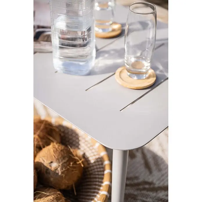 Garden Impressions Loungemöbel-Set Morella - 4 -tlg., Aluminium, Weiß - Heller grauer Outdoor-Tisch mit linearen Schlitzen, mit Wasserflasche und Glas, neben einem Korb Kokosnüsse.