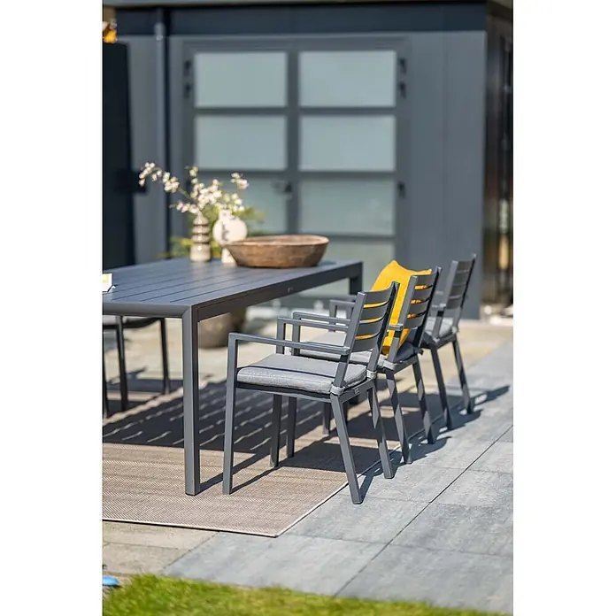 Garden Impressions Stapelsessel Oklahoma - Aluminium, Stapelbar, Dunkelgrau, B x T x H: 56,5 x 55 x 81 cm - Graues Aluminium-Garten-Essgruppe mit Tisch und Stühlen, grauen Polstern und einem gelben Kissen auf einer Terrasse.