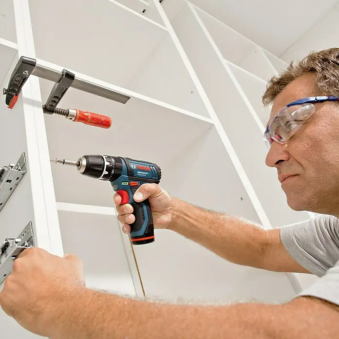 Bosch Professional Akku-Schlagbohrschrauber GSB 12V-15 - 12 V, Ohne Akku, 30 Nm, Ohne Akku, in Karton - Mann verwendet einen Bosch Akku-Bohrschrauber, um eine Halterung an einem weißen Regal zu befestigen. Er trägt eine Schutzbrille.
