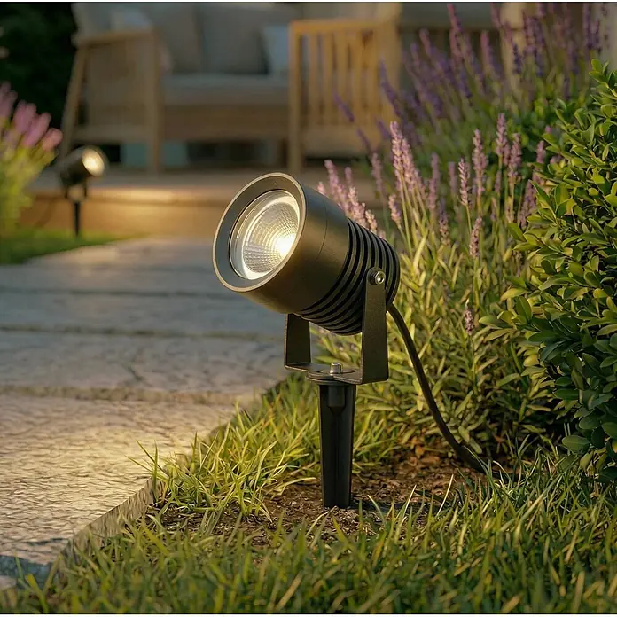 Lucande Erdspießleuchte Irina -  - Schwarzer Outdoor-Strahler mit Erdspieß, beleuchtet einen Gartenweg zwischen Gras und Lavendel.