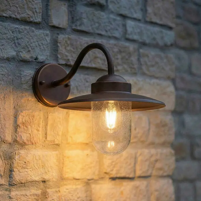 Lindby Außenwandleuchte Bilma - 39,5 x 27,5 x 29 cm, braun / rost, IP44 - Dunkle bronzefarbene Wandleuchte mit strukturiertem Glas, beleuchtet an einer Steinwand.