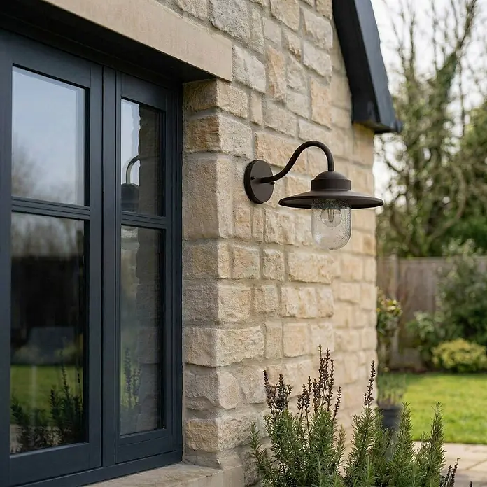 Lindby Außenwandleuchte Bilma - 39,5 x 27,5 x 29 cm, braun / rost, IP44 - Dunkle Metall-Außenwandleuchte mit strukturiertem Glasschirm, an einer Steinwand neben einem dunklen Fenster montiert.