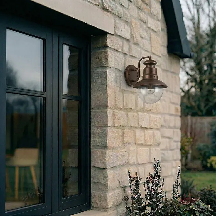 Lindby Außenwandleuchte Eddie - braun / rost, IP44 - Bronze-farbene Außenwandleuchte mit klarer Glaskugel an einer Steinmauer neben einem dunklen Fenster.