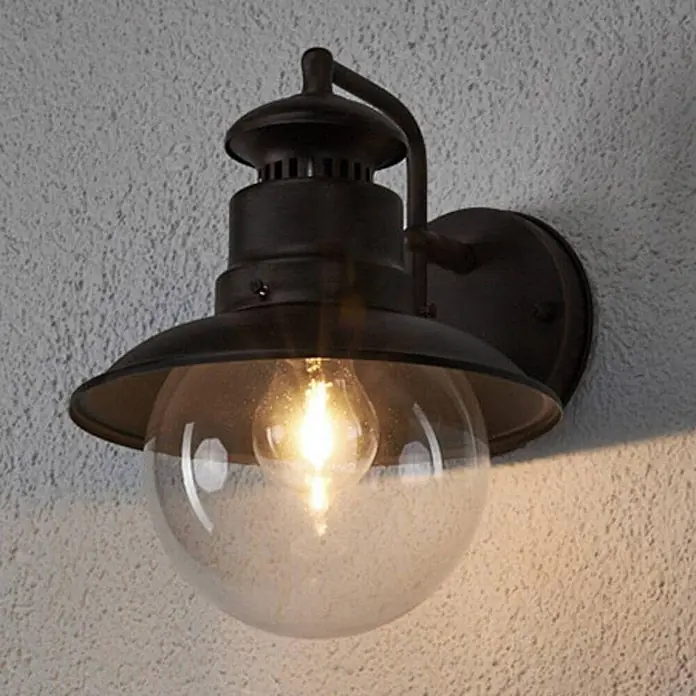 Lindby Außenwandleuchte Eddie - braun / rost, IP44 - Dunkle Bronze Außenwandleuchte mit klarer Glaskugel und leuchtender Filamentbirne an einer strukturierten Wand.