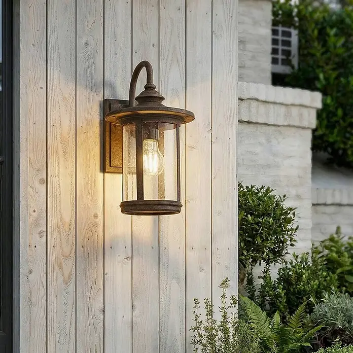 Lindby Außenwandleuchte Zayn - braun / rost, braun / rost, IP23 - Eine wettergegerbte Wandlaterne aus Bronze mit strukturiertem Glas und leuchtender Edison-Glühbirne, an heller Holzverkleidung montiert.
