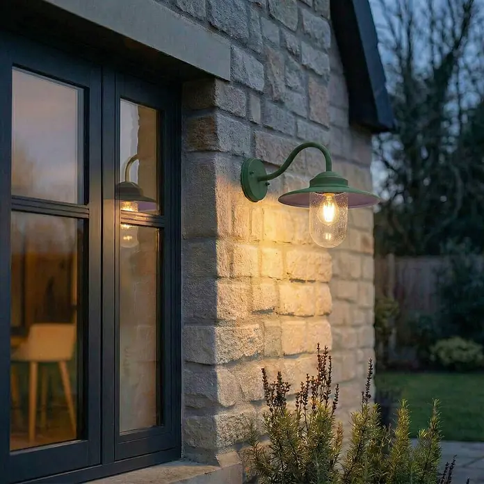 Lindby Außenwandleuchte Bilma - grün, IP44 - Grüne Außenwandleuchte mit strukturiertem Glas, beleuchtet eine Steinwand neben einem Fenster in der Dämmerung.
