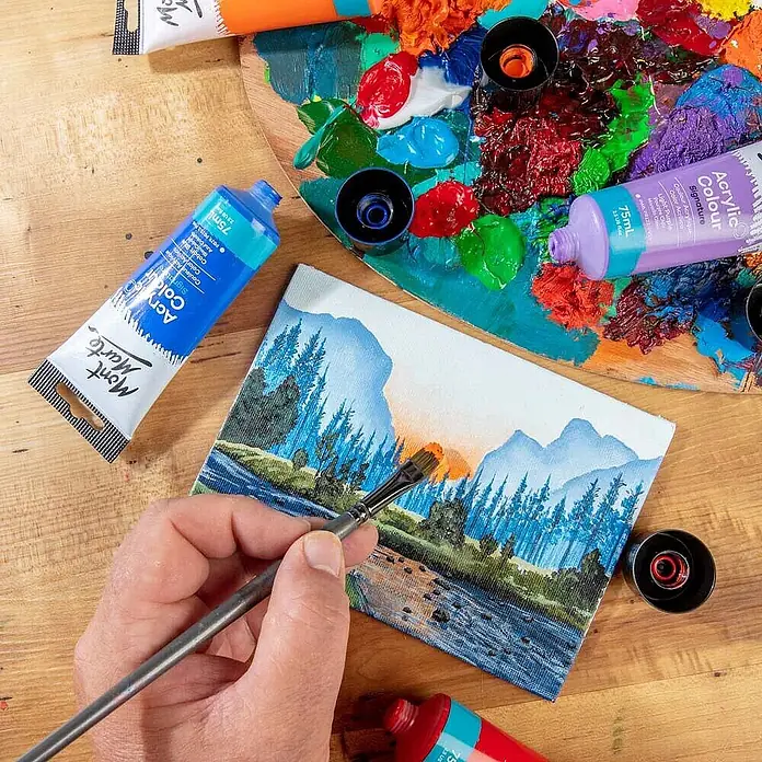 Mont Marte Acrylfarbe Signature - Brillantrot, 75 ml, Tube - Mont Marte Acrylfarben in blauen und lila Tuben, eine Palette mit verschiedenen Farben und eine Hand, die eine Landschaft auf Leinwand malt.