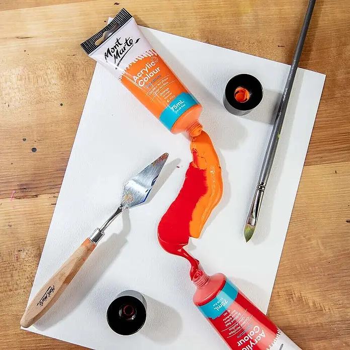 Mont Marte Acrylfarbe Signature - Brillantrot, 75 ml, Tube - Zwei Mont Marte Acrylfarbtuben, eine orange und eine rote, auf eine weiße Oberfläche gedrückt, zusammen mit einem Spachtel und Pinsel.