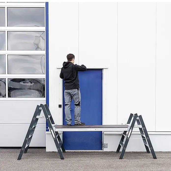 Munk Montagegerüst Einhängeplattform für Klapptritte 1,80x0,48m -  - Mann steht auf einer dunklen Arbeitsplattform, gestützt von zwei anthrazitfarbenen Stehleitern, und arbeitet an einer blauen Tür.