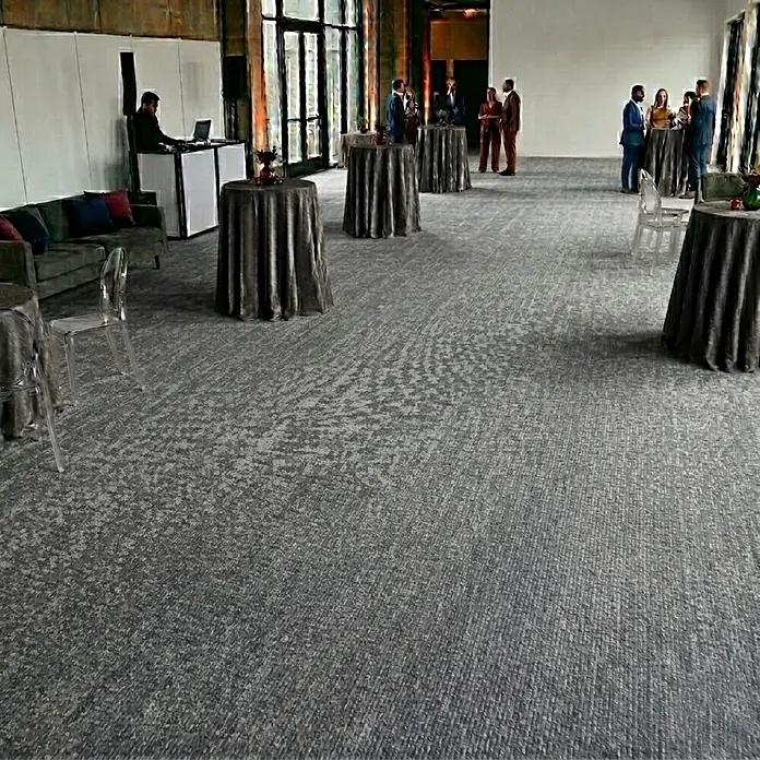 Floordirekt Teppichboden Meterware Sintra - für Messe & Events - Grau meliert - 100 x 350 cm - Breite: 100 cm, Flachgewebe, Polypropylen, Grau meliert - Gemusterter grauer Teppich bedeckt den Boden eines großen Veranstaltungsraums mit Tischen und Gästen.