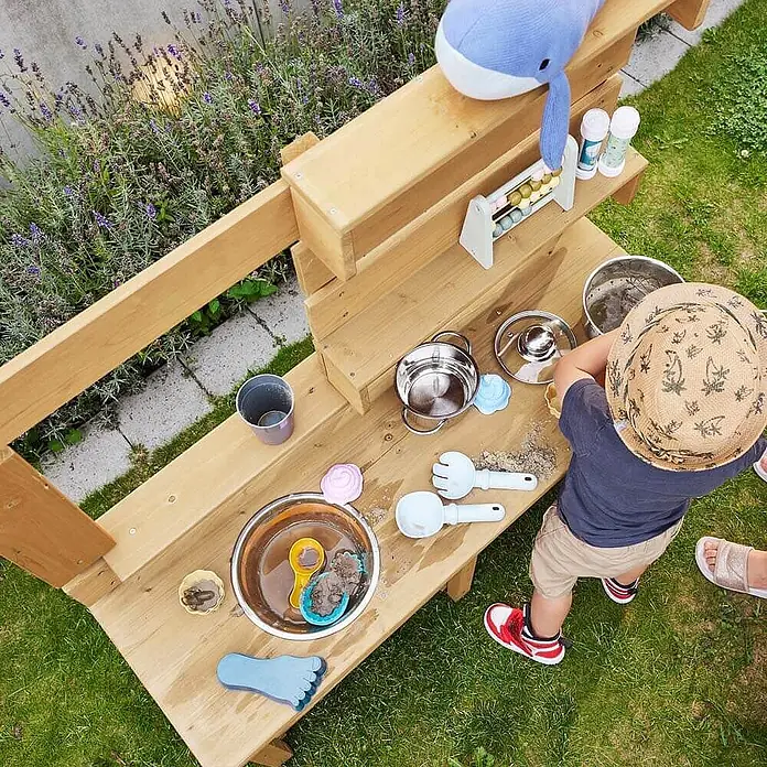 Home Deluxe Matschküche MANDU - Holz, Natur - Hölzerne Matschküche, an der ein Kind draußen mit Töpfen und Sandformen spielt.