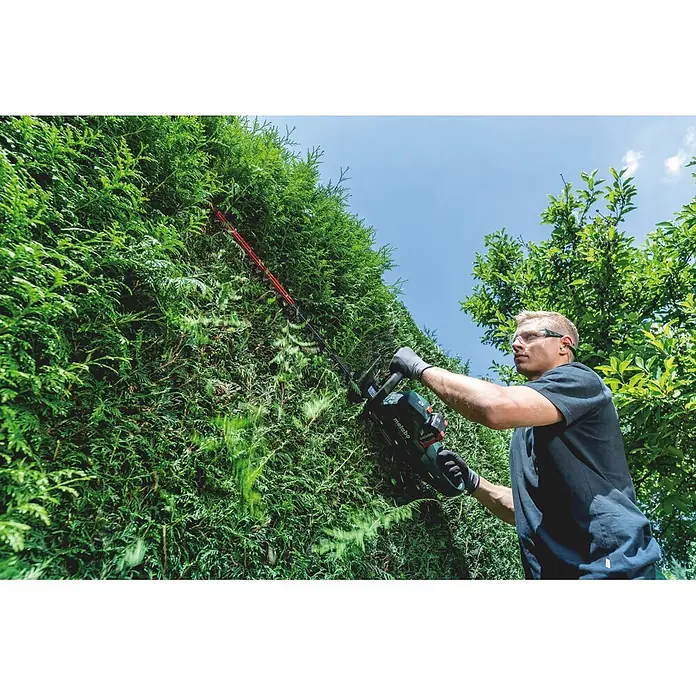 Metabo CAS 18V Akku-Heckenschere HS 18 LTX BL 75 - 18 V, Ohne Akku, Schnittlänge: 70 cm - Mann trimmt Hecke mit grüner/schwarzer Metabo Akku-Heckenschere mit roter Klinge, trägt Schutzbrille und Handschuhe.
