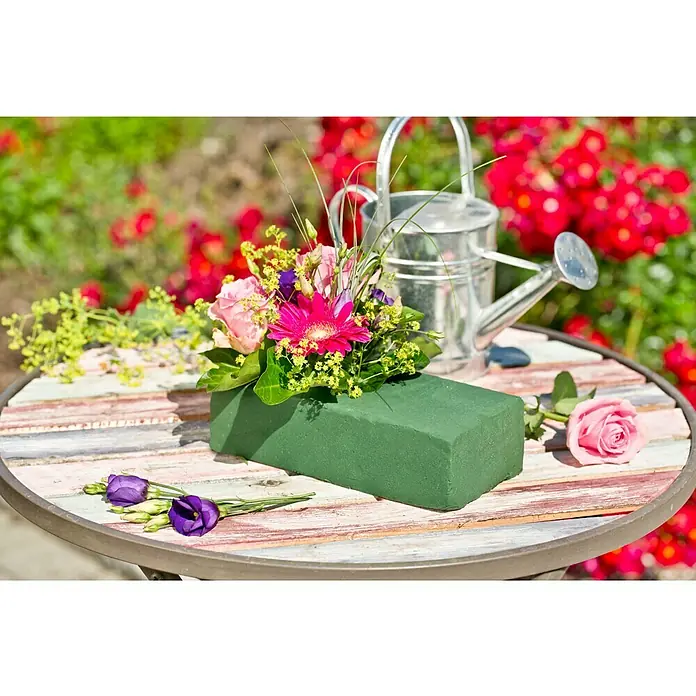 Gardol Steckmasse - 22,5 x 10,5 x 6,5 cm, Geeignet für: Frischblumen - Blumengesteck mit frischen Blumen in einem grünen Steckschaum auf einem Tisch, mit einer Gießkanne und Blumen im Hintergrund.