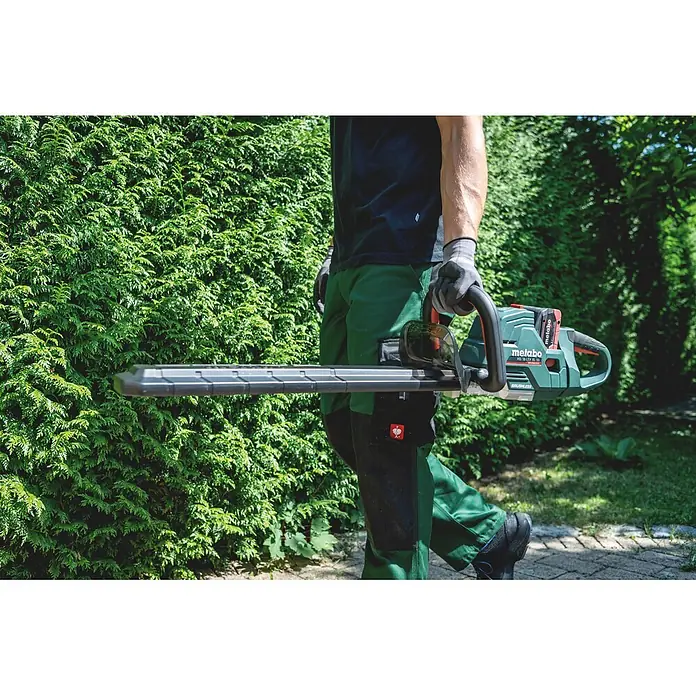 Metabo CAS 18V Akku-Heckenschere HS 18 LTX BL 75 - 18 V, Ohne Akku, Schnittlänge: 70 cm - Eine Person hält eine grün-schwarze Metabo Heckenschere, bereit zum Trimmen einer grünen Hecke.