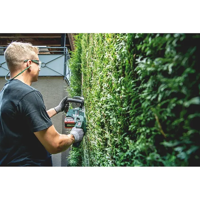 Metabo CAS 18V Akku-Heckenschere HS 18 LTX BL 75 - 18 V, Ohne Akku, Schnittlänge: 70 cm - Person in Schutzbrille und Handschuhen trimmt eine Hecke mit einer grünen Metabo Akku-Heckenschere.