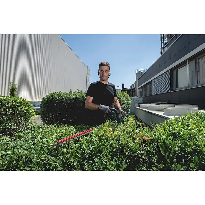 Metabo CAS 18V Akku-Heckenschere HS 18 LTX BL 55 - 18 V, Schnittlänge: 50 cm - Mann trimmt eine grüne Hecke mit einer grün-schwarzen Heckenschere, trägt Schutzbrille und Handschuhe.