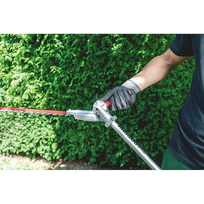 Metabo Heckenscheren-Aufsatz - Passend für: Akku-Multifunktionsantrieb MA 36-18 LTX BL Q - Person mit Handschuhen trimmt eine grüne Hecke mit einem Langstiel-Heckenschneider mit silbernem Schaft und rotem Blatt.