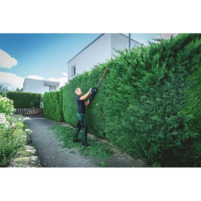 Metabo CAS 18V Akku-Heckenschere HS 18 LTX BL 65 - 18 V, Ohne Akku, Schnittlänge: 60 cm - Mann in grün-schwarzer Arbeitskleidung schneidet eine große grüne Hecke mit einer schwarz-grünen Heckenschere mit roten Klingen.