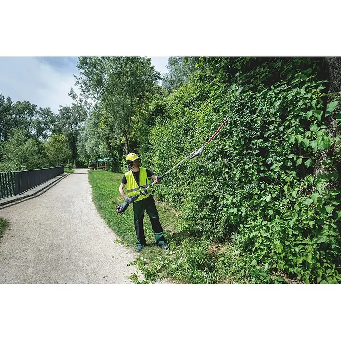 Metabo Heckenscheren-Aufsatz - Passend für: Akku-Multifunktionsantrieb MA 36-18 LTX BL Q - Person in Schutzkleidung schneidet mit einer rot-silbernen Heckenschere mit langem Stiel eine hohe grüne Hecke.