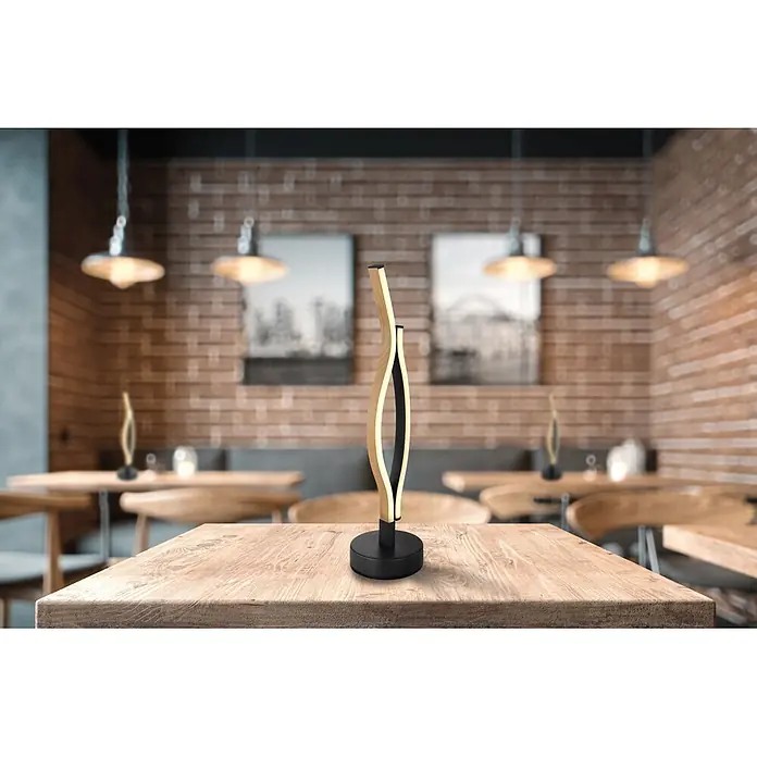 Globo LED-Tischleuchte Geronimo - 12 W, Höhe: 470 mm, Warmweiß - Moderne, wellenförmige Tischlampe aus hellem Holz und Schwarz, die warmes Licht abstrahlt, auf einem Holztisch in einem Café.