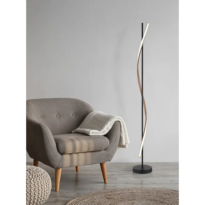 Globo LED-Deckenfluter Watkins - Höhe: 1.507 mm - Moderne Stehleuchte mit schwarzem Ständer und beleuchtetem, wellenförmigem Holz-Lichtstreifen, neben einem grauen Sessel.