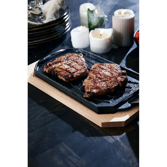 Cozze Grillpfanne - L x B: 30 x 15 cm, Höhe: 1 cm, Gusseisen - Zwei gegrillte Steaks auf einer schwarzen Gusseisenplatte, auf einem hellen Holzbrett serviert, mit aufsteigendem Dampf.