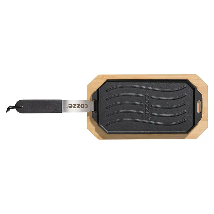 Cozze Grillpfanne - L x B: 30 x 15 cm, Höhe: 1 cm, Gusseisen - COZZE Pizzaschieber mit schwarzem Griff & Metallklinge, in rechteckige COZZE Gusseisen-Grillplatte auf Bambusservierbrett gesteckt.