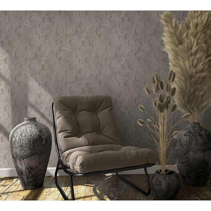Marburg Loft Superior Vliestapete Grafik - Grau-Braun, Grafisch, 10,05 x 0,53 m - Brauner Stoff-Loungesessel mit schwarzem Metallgestell, vor einer grauen sechseckigen Wand, mit Deko-Vasen und Trockenpflanzen auf Holzboden.