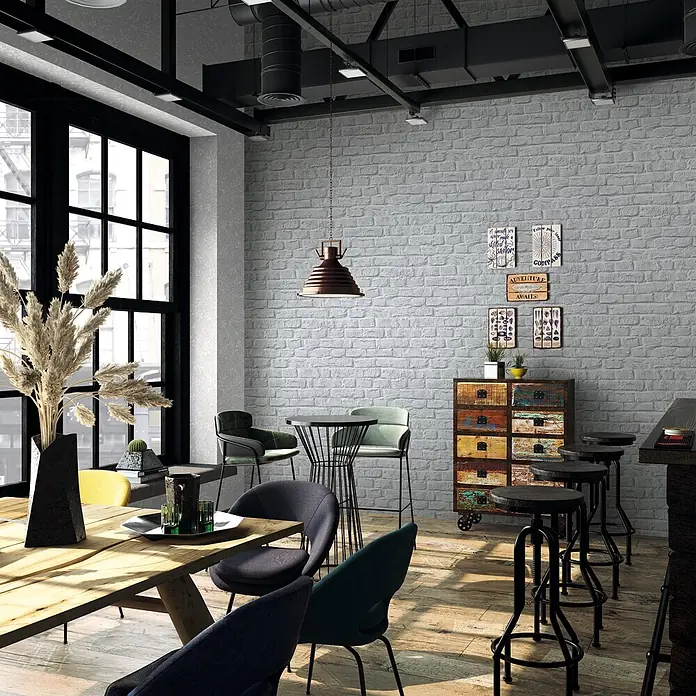 Marburg Loft Superior Vliestapete Steinoptik - Grau, Steinoptik, 10,05 x 0,53 m - Hellgraue Ziegelwand in einem Interieur im Industriestil mit sichtbaren Deckenbalken, großen Fenstern und verschiedenen Möbelstücken.