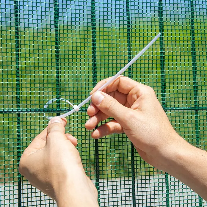 Windhager Drahtgitter - Grün, B x H: 3 x 1 m, Maschenweite: 10 x 10 mm - Hände befestigen einen weißen Kunststoff-Kabelbinder an einem grünen Maschendrahtzaun.
