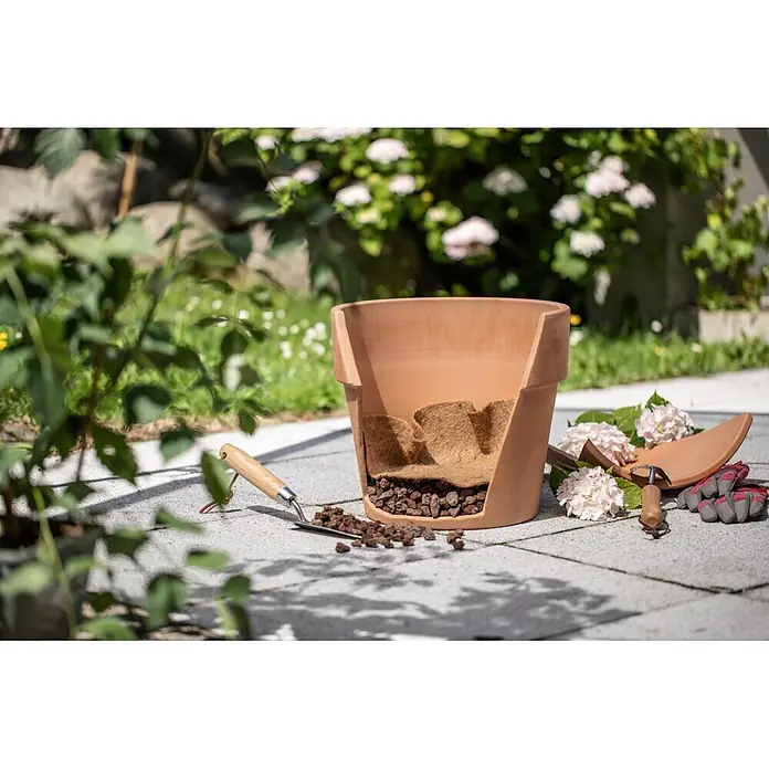 Windhager Trenn- & Filtervlies für Töpfe - 1 x 1 m, Jute - Terrakottatopf, aufgeschnitten, um innere Drainageschichten aus Lavagestein und Kokosfaser zu zeigen, mit Gartengeräten auf einer Terrasse.