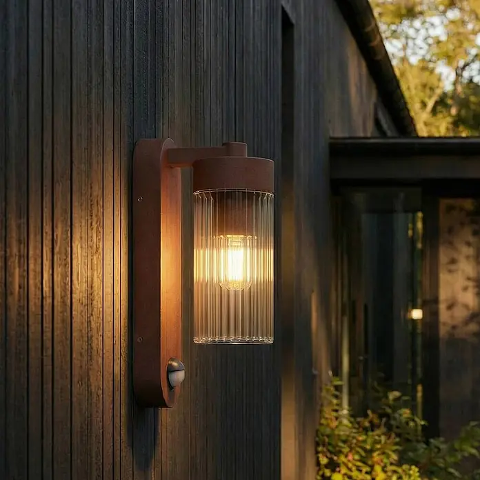 Lindby Außenwandleuchte Kaliro - braun / rost, klar / transparent, IP54 - Rostfarbene Außenwandleuchte mit geriffeltem Glas und sichtbarer Glühbirne, an dunklem Holz befestigt, beleuchtet. Mit Bewegungssensor.