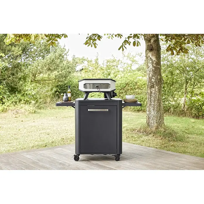 Cozze Unterschrank - L x B x H: 60 x 124 x 83 cm, Schwarz - Cozze Pizzaofen in Weiß und Grau auf einem dunkelgrauen Rollwagen mit Beistelltischen, draußen auf einer Holzterrasse mit Bäumen.