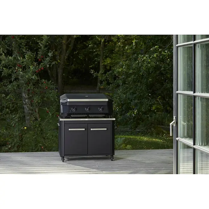 Cozze Outdoorküchen-Modul niedrig - Schwarz/Silber, B x T x H: 93,5 x 67 x 70 cm - Schwarzer Gas-Plancha-Grill mit silbernen Akzenten auf passendem Wagen auf Holzterrasse.