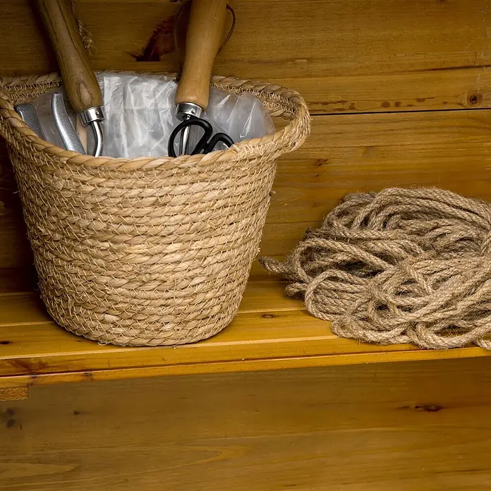 Outsunny Geräteschrank - Außenbreite inkl. Dachüberstand: 79 cm, Außentiefe inkl. Dachüberstand: 49 cm, Naturholz - Gartengeräte mit Holzgriffen in einem geflochtenen Korb, neben einem Seilknäuel, auf einem Holzregal.