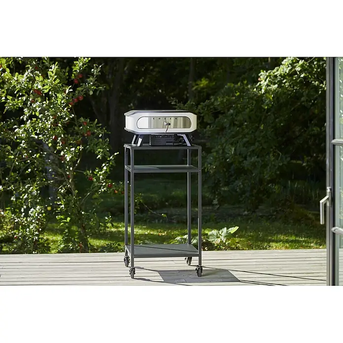 Cozze Pizzaofen mit drehbarem Pizzastein - Durchmesser Grillfläche: 42,5 cm, 6 kW, Weiß/Schwarz, Stahl - Ein weiß-silberner Outdoor-Pizzaofen auf einem dunkelgrauen Servierwagen auf einer Holzterrasse.