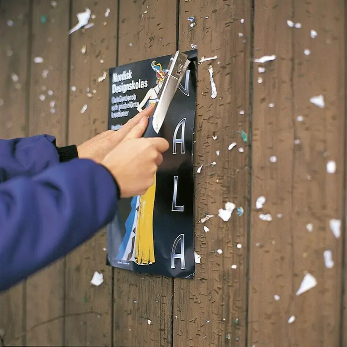 Rapid Hammertacker R19 - Klammertiefe: 4 mm - 6 mm, Geeignet für: Feindrahtklammern Typ 13 (4 - 6 mm), Blisterpack - Nahaufnahme einer Person, die mit einem Tacker ein Poster an einer Holzwand befestigt. Das Poster wirbt für die Nordisk Designskola.