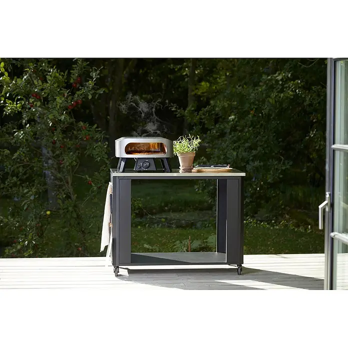 Cozze Outdoorküchen-Modul - Schwarz/Silber, B x T x H: 119 x 60 x 90 cm - Ein silberner Outdoor-Pizzaofen gart eine Pizza mit aufsteigendem Rauch auf einem dunkelgrauen Outdoor-Küchentisch im Garten.