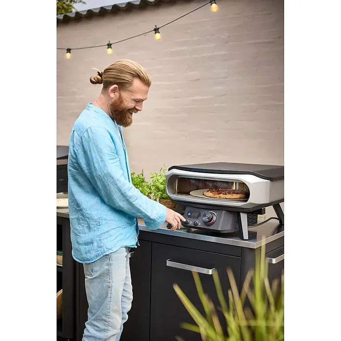 Cozze Pizzaofen Elektro Premium 13" - Grillfläche (B x T): 34 x 34 cm, 2,2 kW - Mann bedient einen dunkelgrauen Cozze Pizzaofen, der eine Pizza auf einer Outdoor-Kücheneinheit zubereitet.